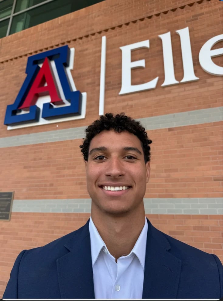 Man in navy blazer and light blue shirt smiling in front of red brick building with Arizona Wildcats logo and partial text visible